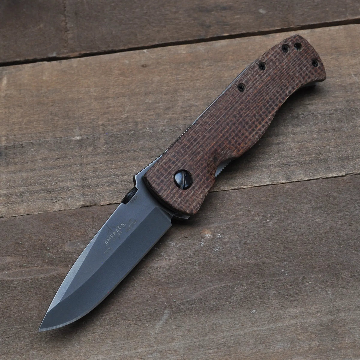 The Emerson Knives, Inc. CQC-7A-NW with Brown Burlap is a limited edition folding pocket knife featuring a dark, partially serrated blade and a textured brown handle, shown open on a wooden surface.