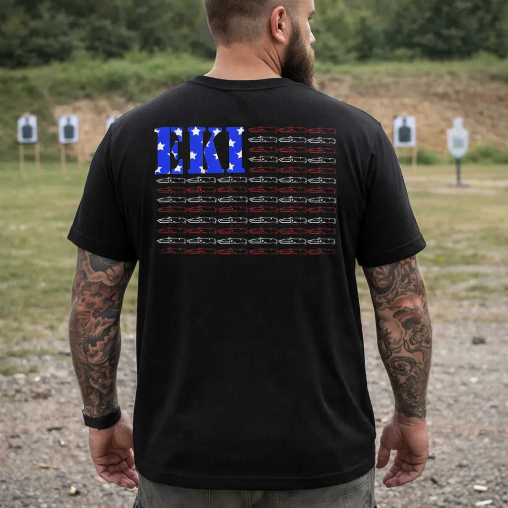A man with tattooed arms stands outdoors, wearing the Emerson Flag T-Shirt by Emerson Knives, Inc. The shirt's back displays an American flag design—EK in blue and white stars replace the original stars, and knives form the stripes. Targets appear in the distance.