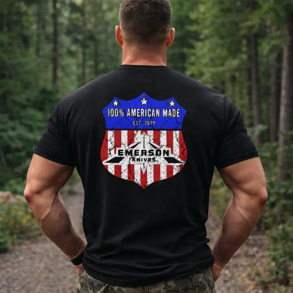 A muscular man stands on a forest path, wearing the Route 66 T-Shirt by Emerson Knives, Inc., featuring a black design with a red, white, and blue shield and "100% American Made Est. 1979" on the back.