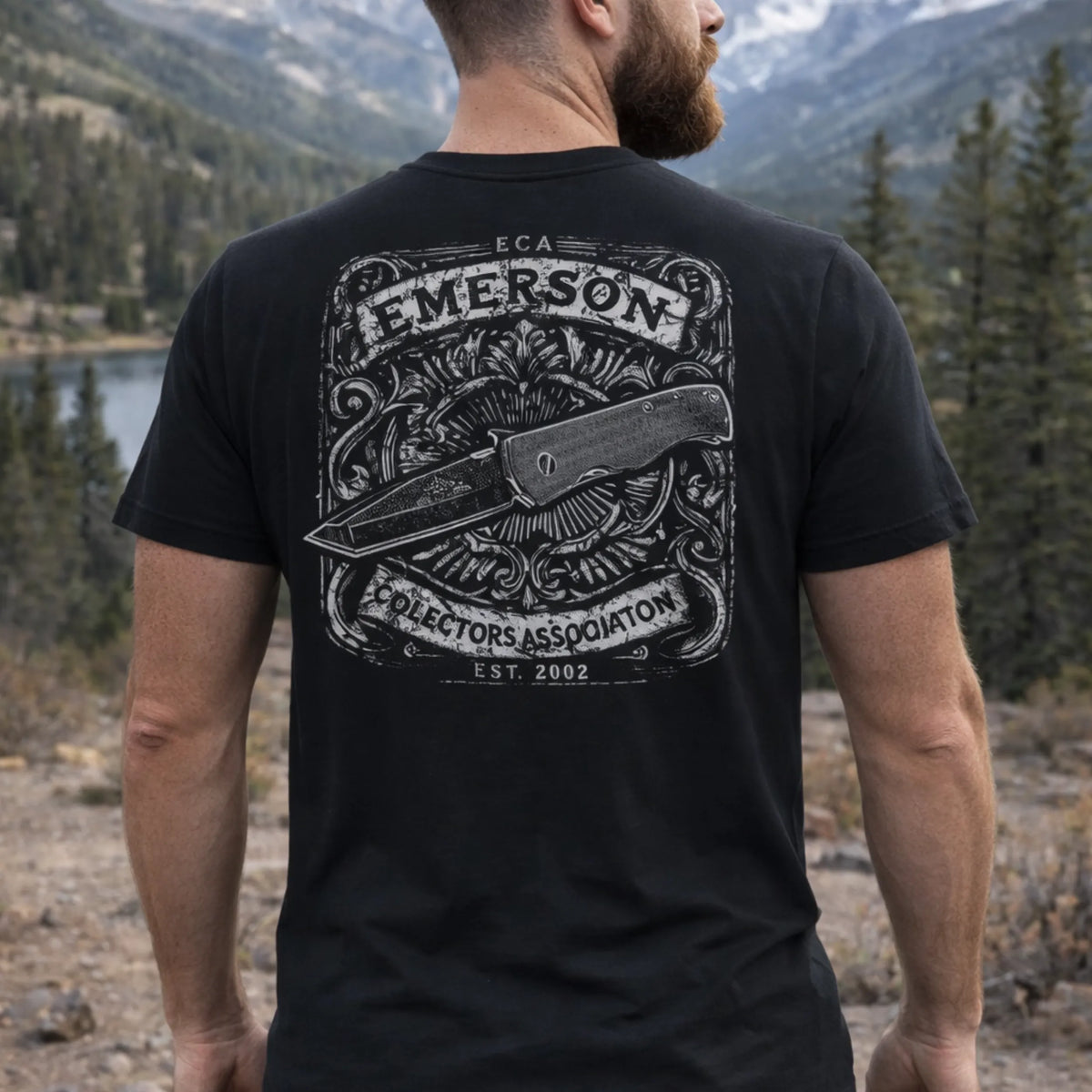 A man stands by a lake and trees, wearing the Emerson Knives, Inc. ECA T-Shirt in black, which features an ornate folding knife design and the words "Emerson Collectors Association Est. 2002" on the back.