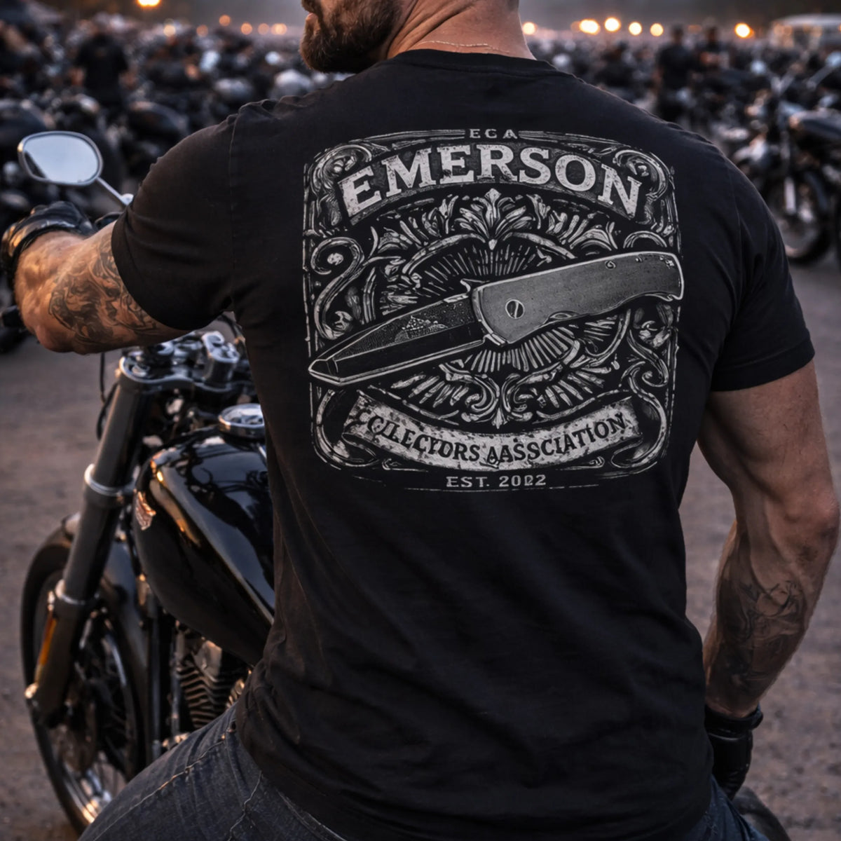 A man on a motorcycle wears the ECA T-Shirt by Emerson Knives, Inc., featuring an ornate folding knife design and "Emerson Collectors Association Est. 2002" text. Motorcycles and people can be seen in the background.