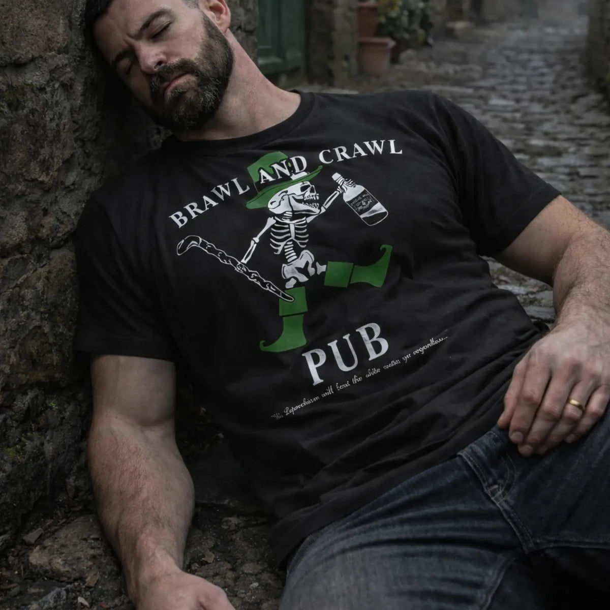 A bearded man in an Emerson Knives, Inc. Brawl and Crawl Pub T-Shirt with a quirky slogan and skeleton design relaxes against a stone wall on a cobblestone street, eyes closed, one hand on his knee, as if murmuring “Slainte” in his sleep.