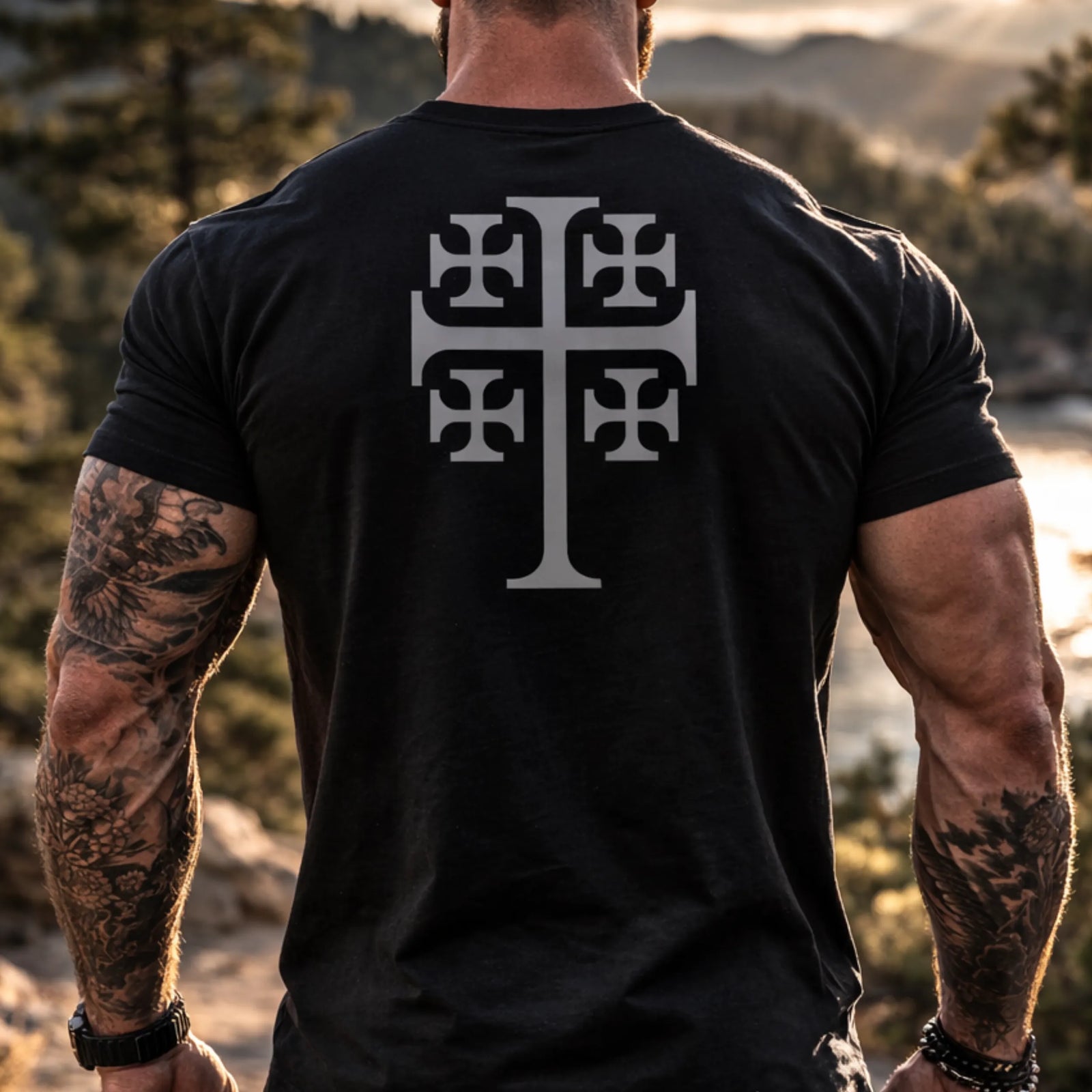A muscular person with tattooed arms stands outdoors wearing the Emerson Knives, Inc. Deus Vult T-Shirt, which features a large Jerusalem Cross and "Deus Vult" on the back. Trees and a river are blurred in the background.