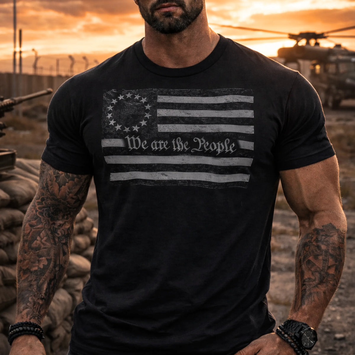 At sunset outdoors, a muscular man with tattooed arms wears the Emerson Knives, Inc. We Are the People T-Shirt featuring a distressed Betsy Ross flag and “We the People.” Military helicopters and sandbags are seen in the background.