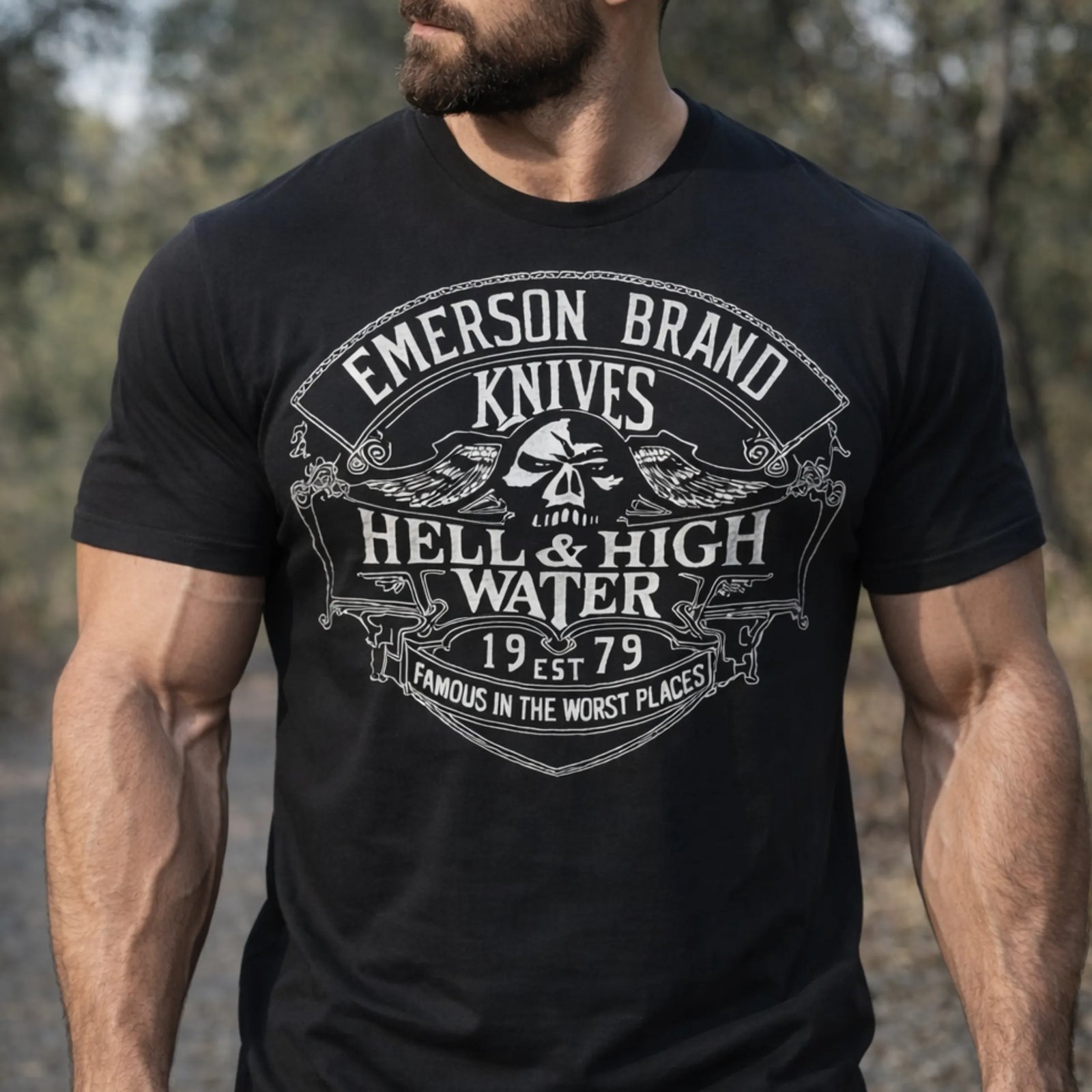 A bearded man stands outdoors wearing the Emerson Knives, Inc. Hell and High Water T-Shirt, featuring a skull with crossed knives and bold "Famous in the Worst Places" text. No product description is provided.
