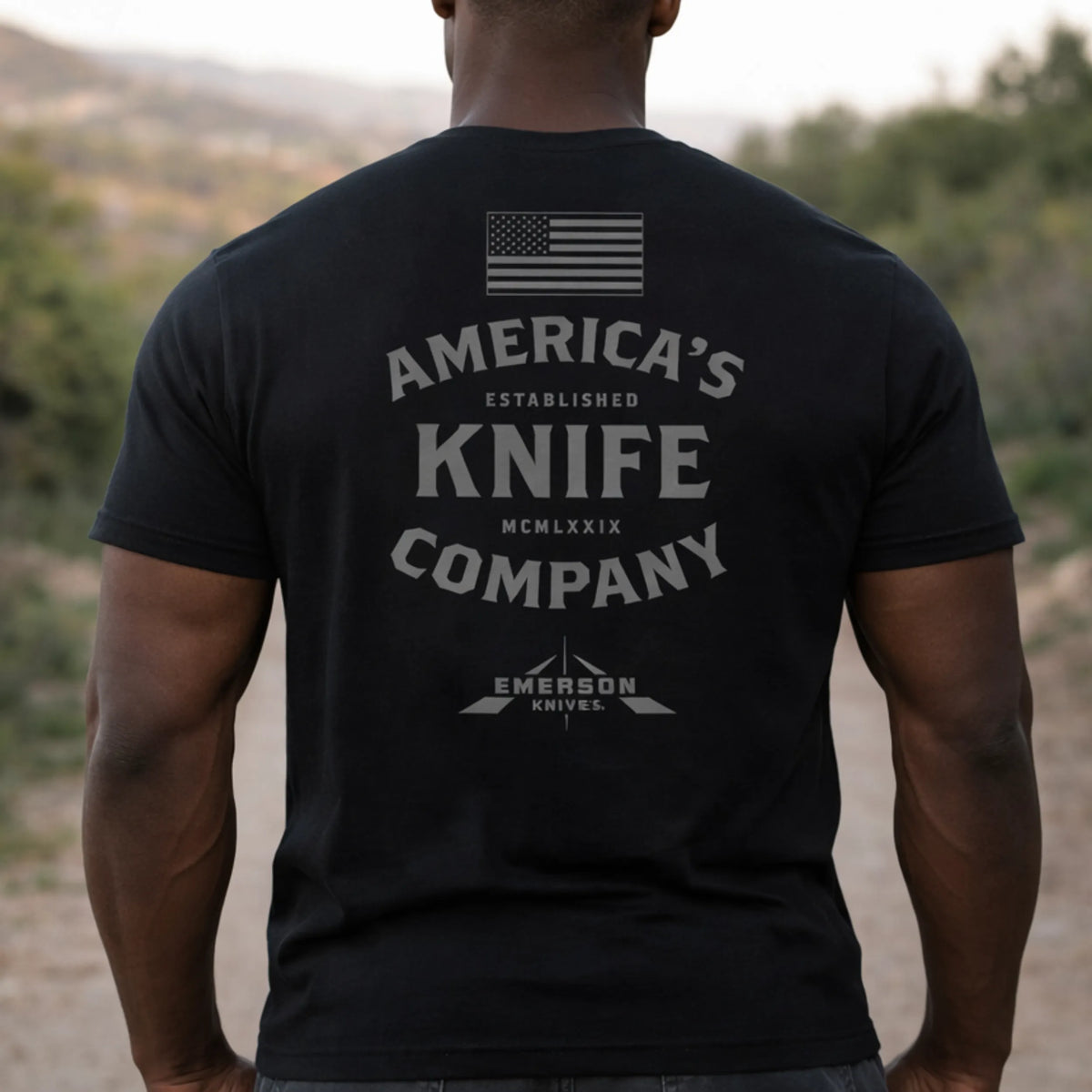 A man stands outdoors wearing the Emerson Knives, Inc. "America's Knife Company T-Shirt," featuring a U.S. flag and company branding on the back, with trees and hills in the background.