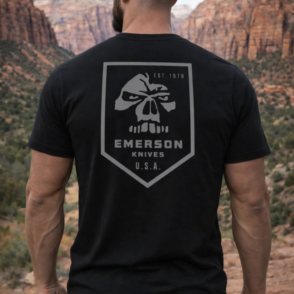 A man stands outdoors in a canyon wearing the Emerson 3.0 T-Shirt by Emerson Knives, Inc., which features a large skull logo and “EMERSON KNIVES U.S.A. EST. 1979” on the back, with rocky cliffs and greenery in the background.