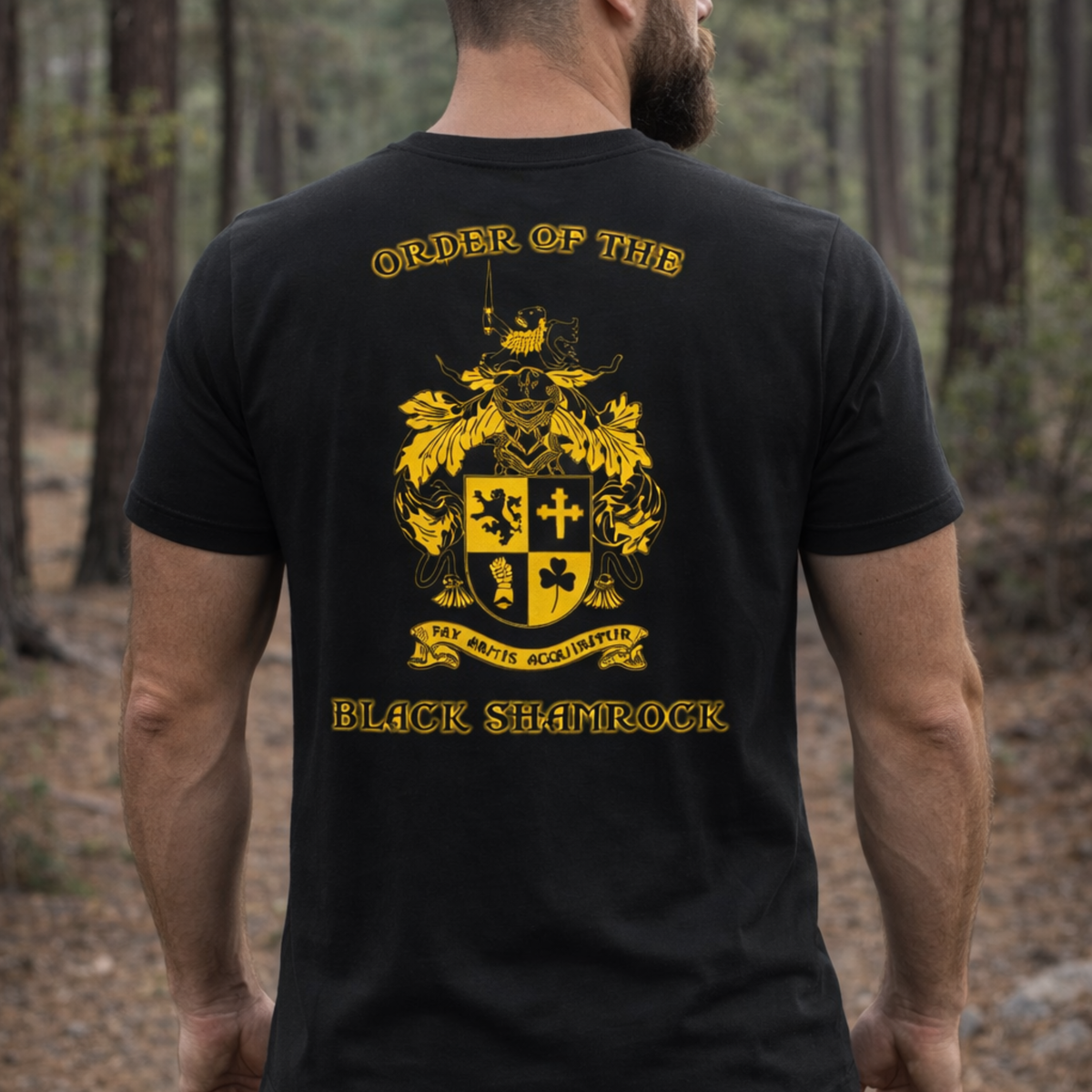 A man stands in a forest wearing the OBS T-Shirt by Emerson Knives, Inc., featuring a gold crest, “Order of the Black Shamrock” text, a Latin motto, and medieval-style symbols on the back.