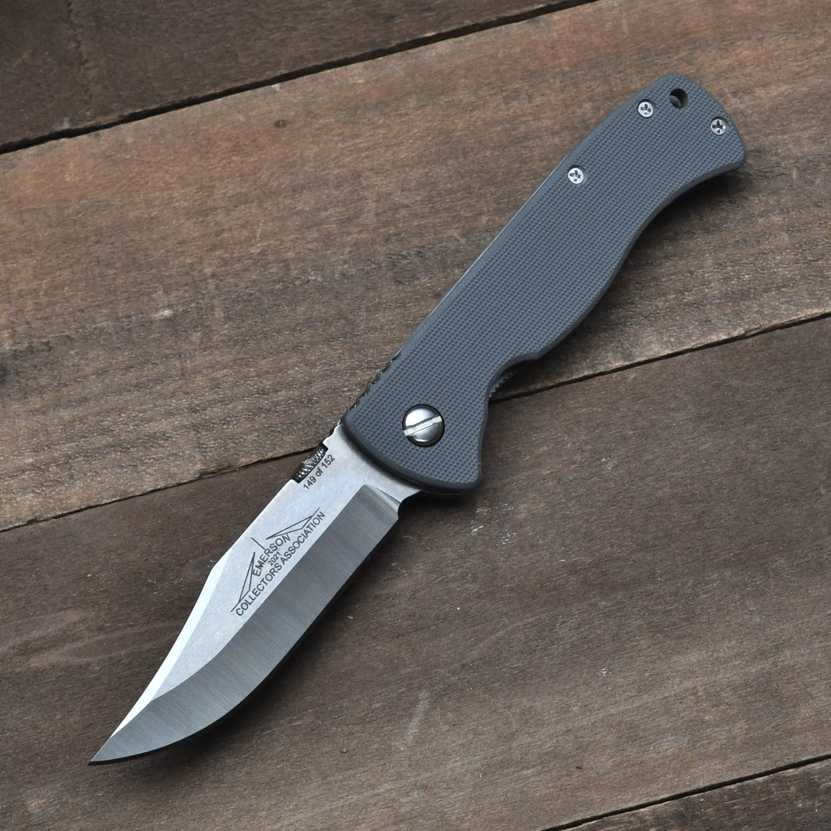 The ECA KOTY EX-100 by Emerson Knives, Inc., a Limited Edition Christmas Knife with a gray textured handle and partially serrated stainless steel blade, is displayed open on a wooden surface.