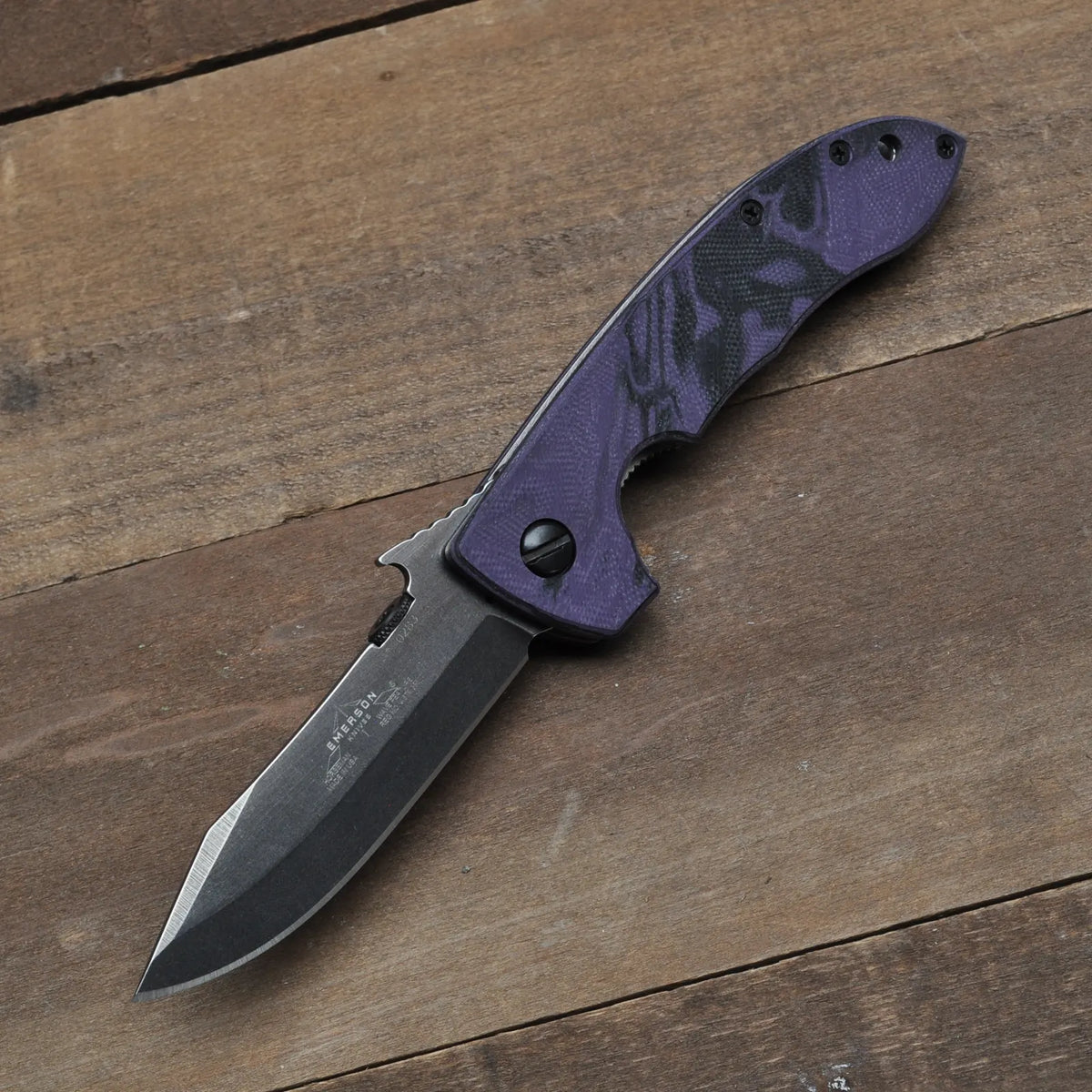 The Emerson Knives, Inc. Horseman Purple Black Handles is a limited edition folding pocket knife with a dark, partially serrated blade and a textured purple handle, displayed on a wooden surface.
