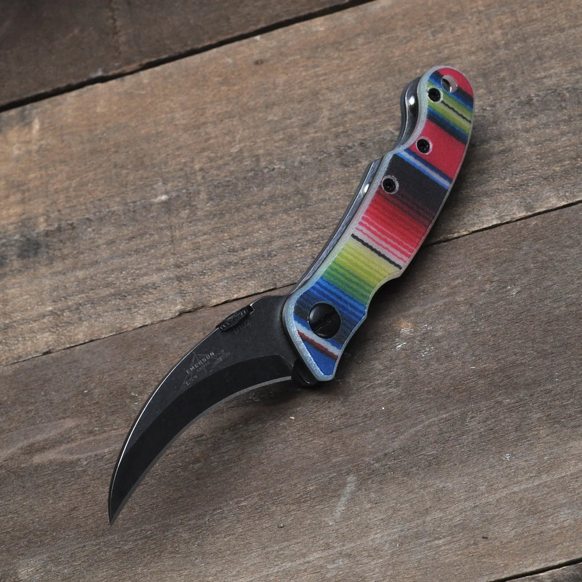 The Emerson Knives, Inc. Mini Elvia Serape TF is a limited edition folding karambit with a curved black blade and a handle accented by colorful horizontal stripes, shown resting on a wooden surface.
