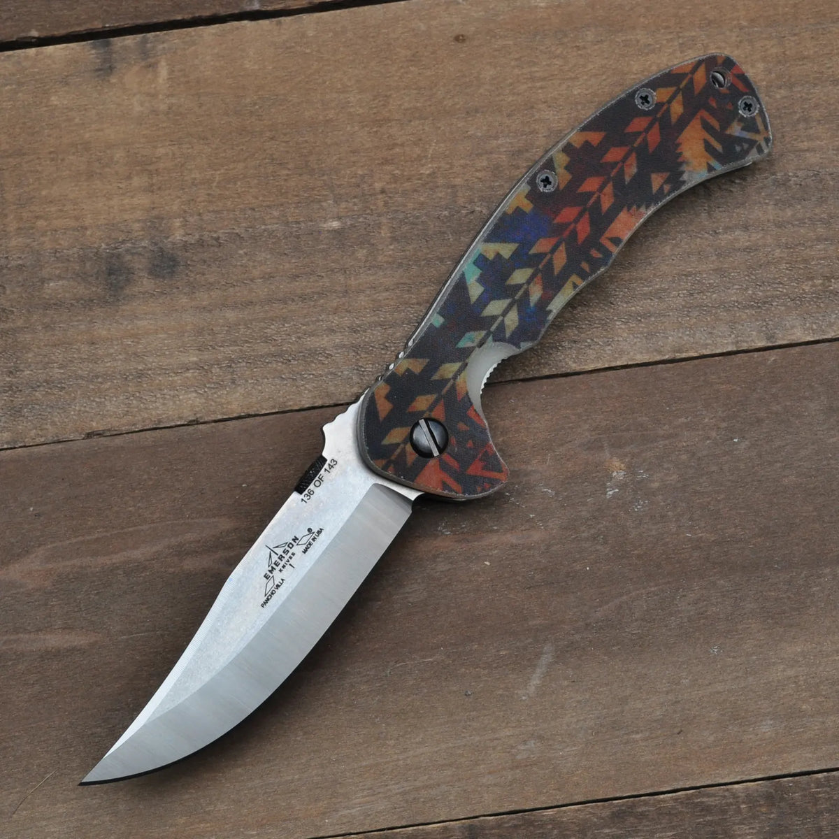 The Emerson Knives, Inc. Pancho Villa Southwestern G-10 folding pocket knife, featuring a geometric multicolored handle, lies open on a wooden surface. This limited edition shiny, slightly curved blade reflects light.