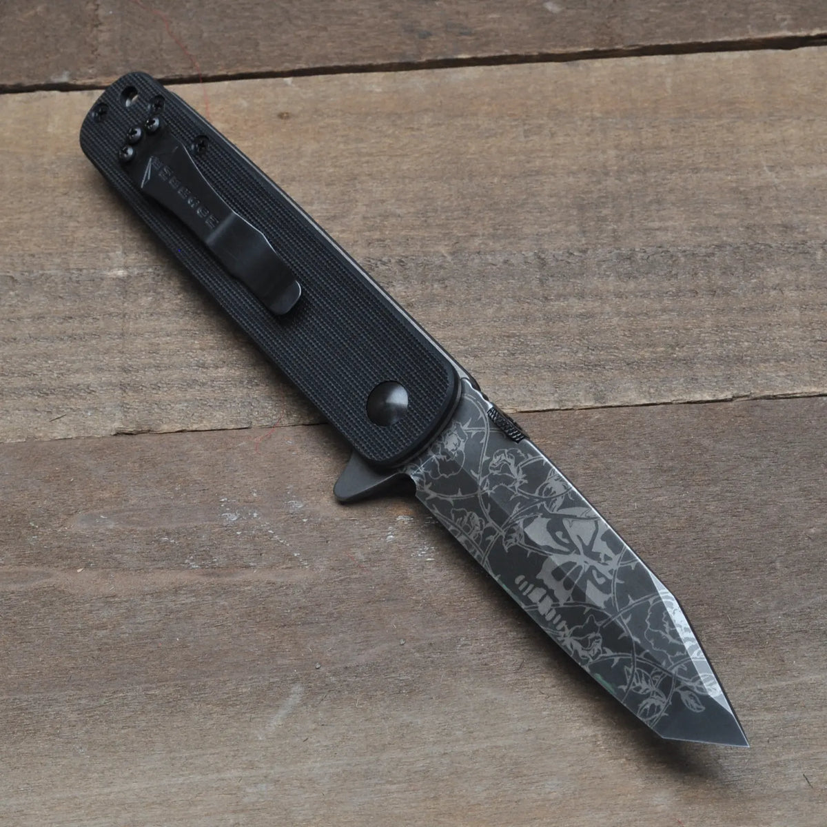 The Emerson Knives, Inc. Tanto A-100 Flipper- Vines, Skull, and Roses Engraving TF is a limited edition folding pocket knife with a black textured handle and an engraved blade featuring skulls, vines, and roses.
