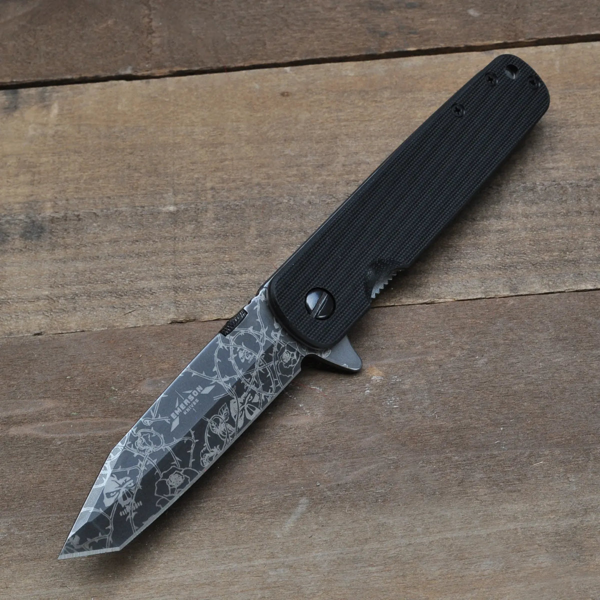 The Emerson Knives, Inc. Tanto A-100 Flipper- Vines, Skull, and Roses Engraving TF folding knife with a black handle and an engraved tanto blade is displayed open on a wooden surface.